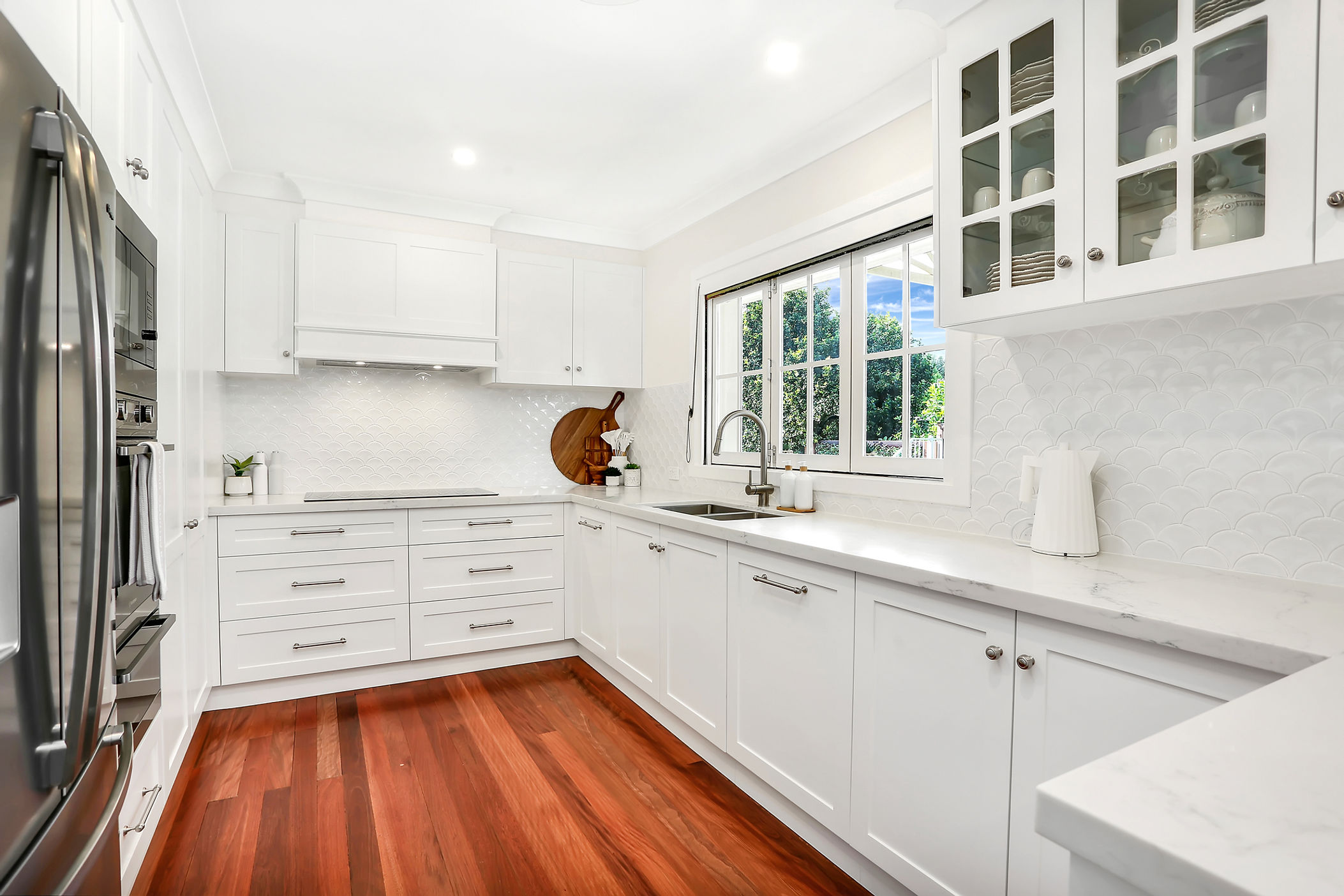 White shaker kitchen with timber floors, scalloped splashback and a window over the sink