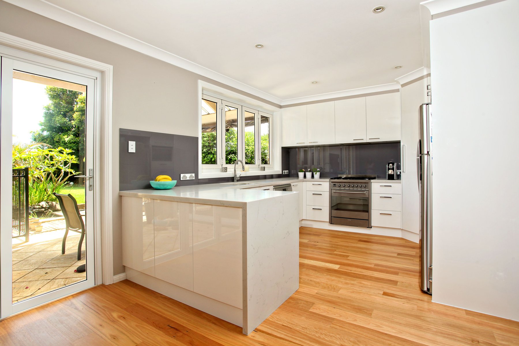 White kitchen island opening to a garden, with timber stools and a stone benchtop
