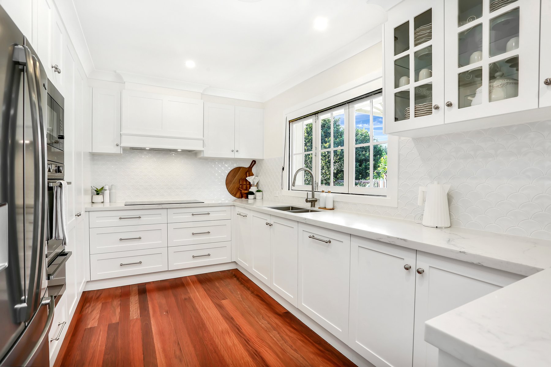 White shaker kitchen with timber floors, a window over the sink and glass-front cabinets